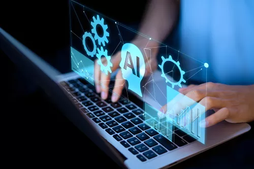 Personne, dont on ne voit pas le visage, qui tape sur le clavier d'un ordinateur portable. A la verticale du clavier, en transparence, AI et roues crant&eacute;es.