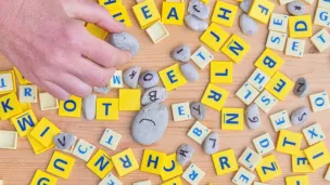 Pions avec des lettres dessus, type Scrabble, blancs et jaunes, petits galets avec des lettres &eacute;crites dessus, tous m&eacute;lang&eacute;s, main sur la gauche saisissant un pion