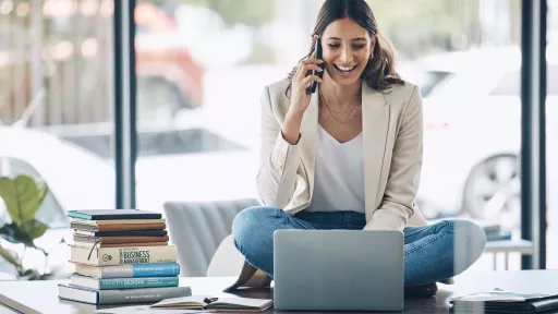 Worker, laptop and phone call laughing while sitting on desk in relax startup, marketing company or business. Happy smile, talking and woman on mobile communication technology, laptop or books study