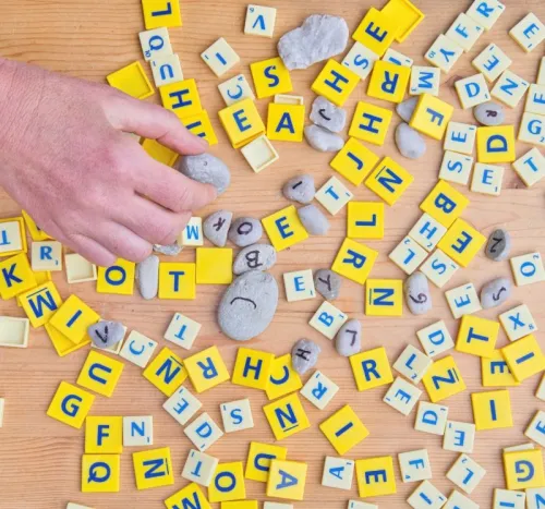 Pions avec des lettres dessus, type Scrabble, blancs et jaunes, petits galets avec des lettres &eacute;crites dessus, tous m&eacute;lang&eacute;s, main sur la gauche saisissant un pion